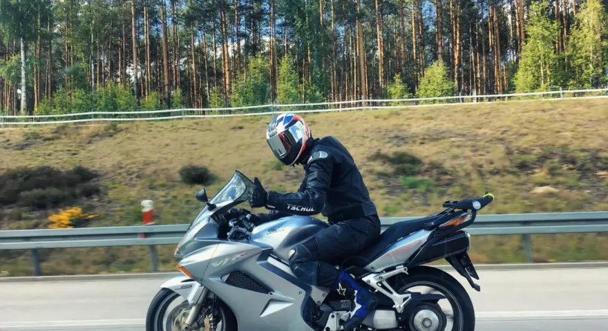 Motocyklista w kasku jedzie na srebrnym Honda VFR 800 wzdłuż leśnej drogi.