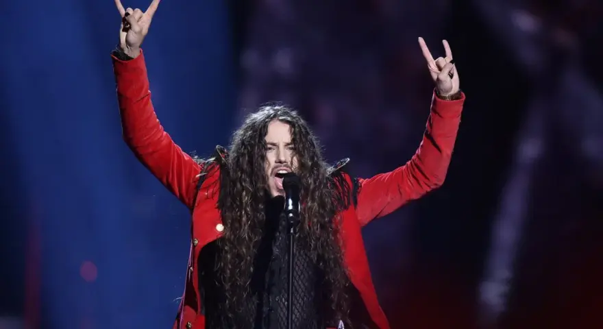 Michał Szpak na Eurowizji w czerwonej marynarce, z uniesionymi rękami i gestem rock'n'rolla.
