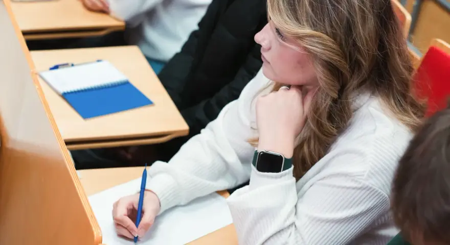 Studentka pisze notatki na wykładzie, opierając podbródek na dłoni.
