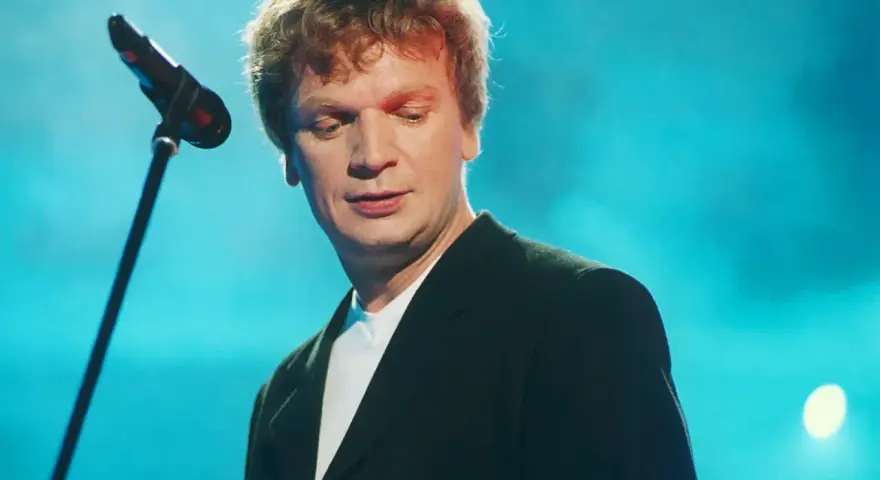 Grzegorz Ciechowski, w czarnym garniturze, pochyla się nad mikrofonem. Jego niezapomniane utwory wciąż brzmią w sercach fanów.