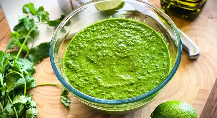 Frisch zubereitetes mojo verde in einer Glasschüssel, umgeben von Koriander, Knoblauch, Limetten und Olivenöl.