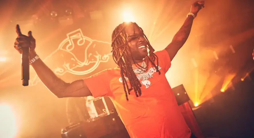 Rapper mit Dreadlocks und orangefarbenem T-Shirt singt mit erhobenen Armen und Mikrofon. Die Bühne ist in warmes Licht getaucht, perfekt für energiegeladene Hip-Hop-Songs.