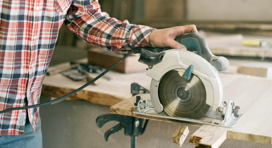 A person in a plaid shirt demonstrates how to use a circular saw, cutting wood with sawdust flying.