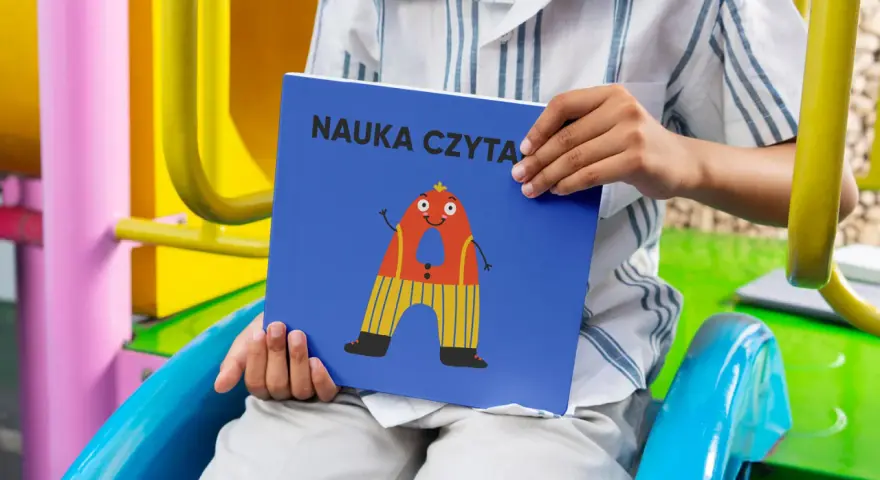 Chłopiec trzyma książkę "Nauka czytania dla 6 latka" z zabawną literką "A" na okładce.