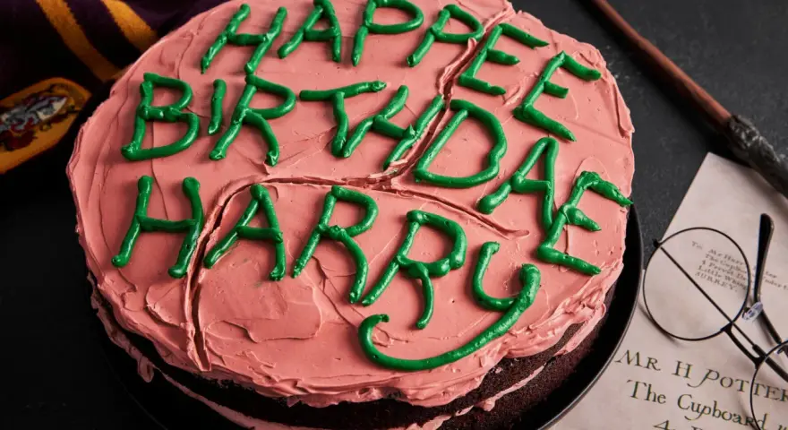 Ein Harry Potter Kuchen mit rosa Zuckerguss und grüner Schrift "Happy Birthday Harry". Daneben liegen ein Schal, eine Brille und ein Brief.