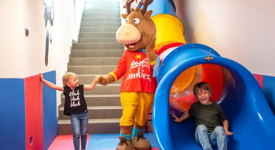 Mascot and two children in a colorful indoor playground with a slide and stairs.