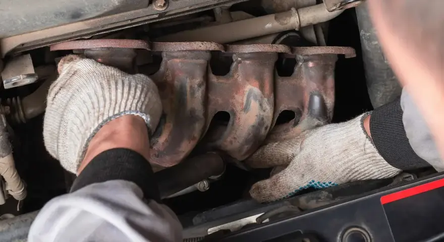 Mechanic's gloved hands work on a rusty exhaust manifold, possibly to fix an engine ticking noise.
