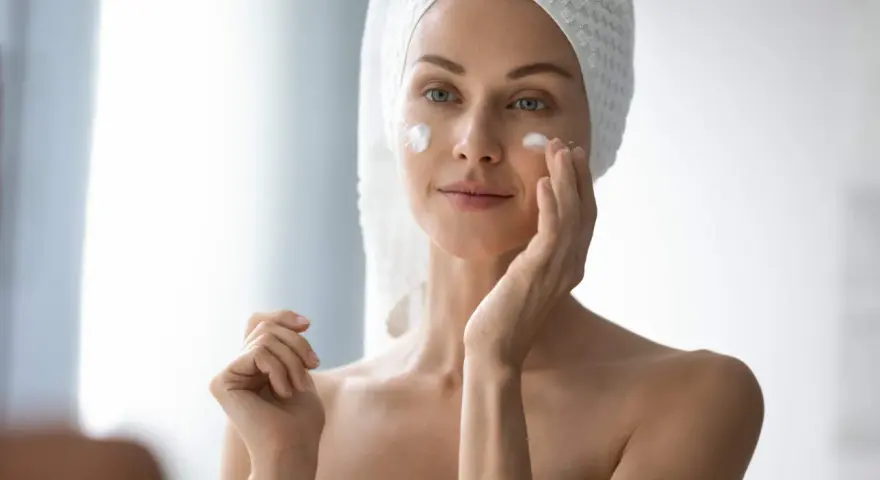Femme appliquant de la crème sur son visage pour lutter contre la xerose cutanée. Elle a une serviette sur la tête.