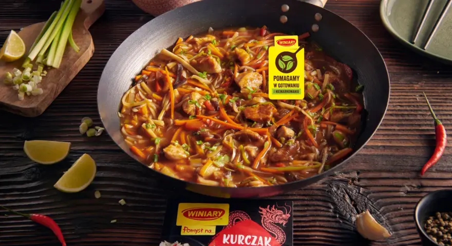 Kurczak w pięciu smakach po chińsku w woku, z warzywami i ryżem w tle. Obok limonki, chili i czosnek.