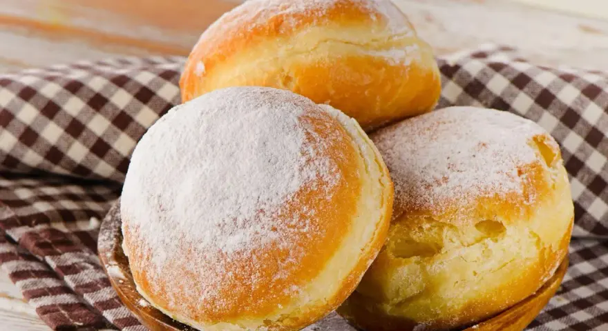 Trzy puszyste pączki, dwa posypane cukrem pudrem, leżą na drewnianej misce. Idealne na tłusty czwartek, te fit pączki kuszą apetycznym wyglądem.
