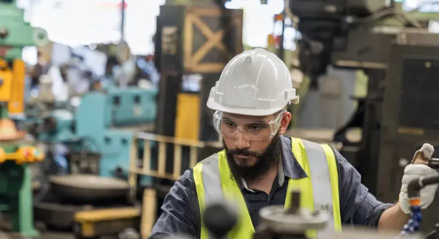 Obrero con casco y gafas de seguridad trabajando en una máquina en una fábrica, parte esencial de la manufactura moderna.