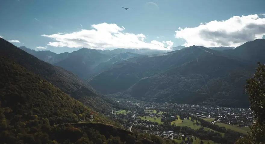 Vue aérienne d'une vallée verdoyante avec un village niché, entouré de montagnes majestueuses. Un planeur plane dans le ciel bleu, rappelant les paysages de Toulouse et ses environs.