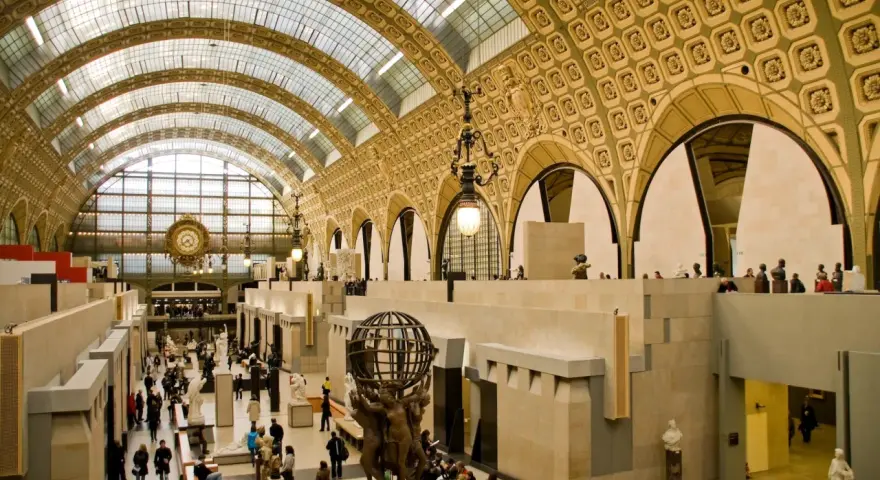 Interior del museo d'Orsay, con su impresionante techo de cristal y arcos dorados. Visitantes pasean entre esculturas y obras de arte.