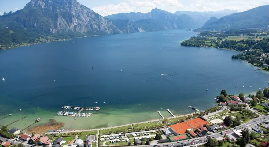 Idyllischer **Camping Traunsee** mit Blick auf den See und die Berge. Boote liegen im Hafen, Tennisplätze und Wohnwagen sind zu sehen.