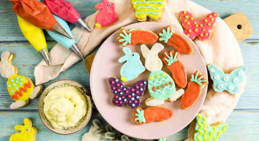 Bunte Osterkekse in Hasen-, Karotten- und Schmetterlingsform, bereit für ein einfaches Osterkekse Rezept. Spritzbeutel mit Zuckerguss liegen bereit.