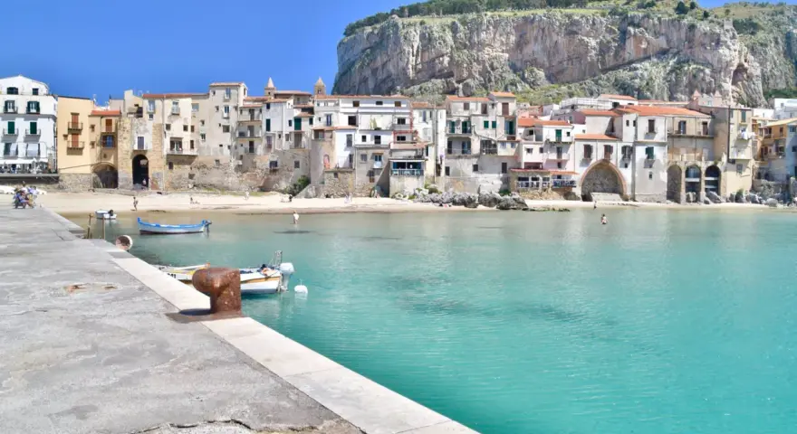 Coastal town with buildings nestled against a cliff, turquoise water, and boats. Perfect for a 10 days in Sicily adventure.