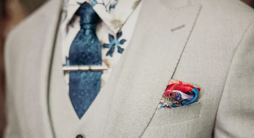 Elegancki mężczyzna w jasnej marynarce, z niebieskim krawatem i spinką do krawata jak nosić. W kieszeni kolorowa poszetka.