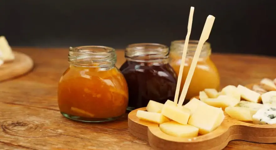 Tre vasetti di marmellata, uno arancione, uno scuro e uno giallo, accompagnano un tagliere di formaggio a cubetti, pronto per essere gustato.