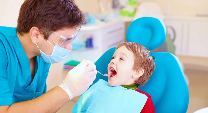 Dentysta bada uśmiechniętego chłopca, sprawdzając, kiedy najpóźniej pierwszy ząbek pojawi się u malucha.