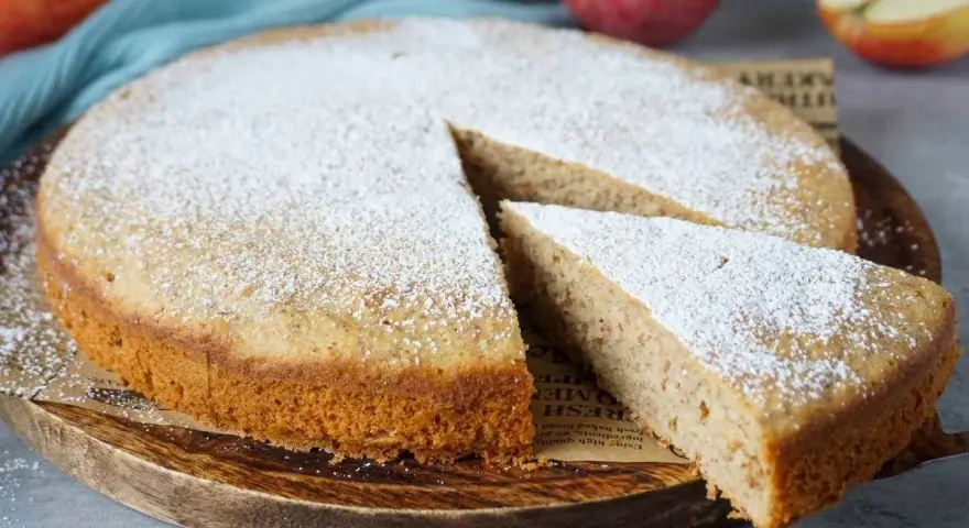 Ein schneller, saftiger Kuchen mit Apfelmus, bestäubt mit Puderzucker. Ein Stück wird gerade herausgenommen.