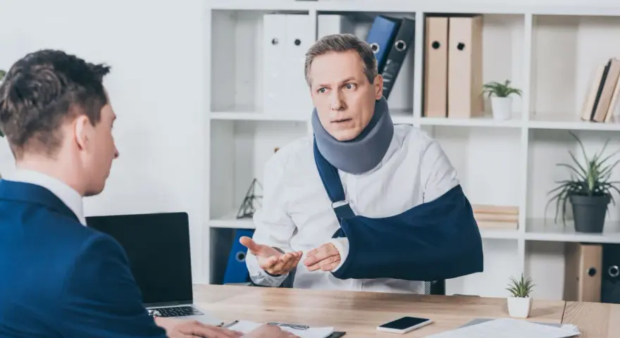 Mężczyzna w kołnierzu ortopedycznym i temblaku rozmawia z drugim mężczyzną przy biurku.