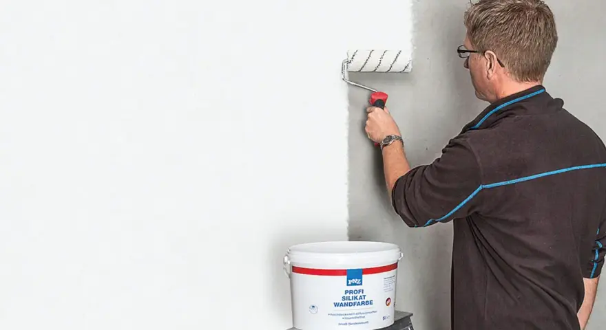 Mann streicht mit einer Farbrolle eine Wand weiß. Der Eimer mit der "Profi Silikat Wandfarbe" steht daneben. Perfekt für das Streichen bei Auszug.
