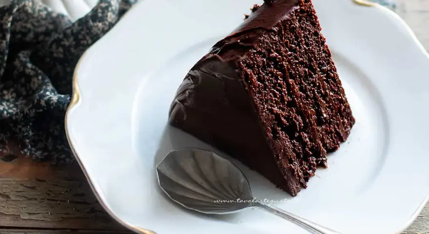 Una fetta di torta al cioccolato morbida e umida, ricoperta di glassa lucida, su un piatto bianco con un cucchiaino.