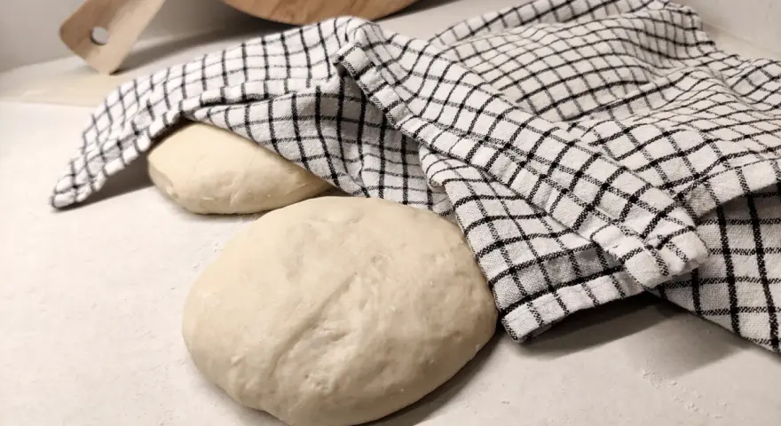 Two balls of pizza dough on a counter, one covered with a checkered cloth, with a wooden pizza peel in the background.