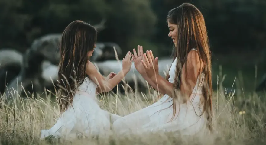 Dwie dziewczynki w białych sukienkach siedzą na łące i bawią się w "pajacyka". To idealna wyliczanka dla dzieci, która rozwija koordynację ruchową.