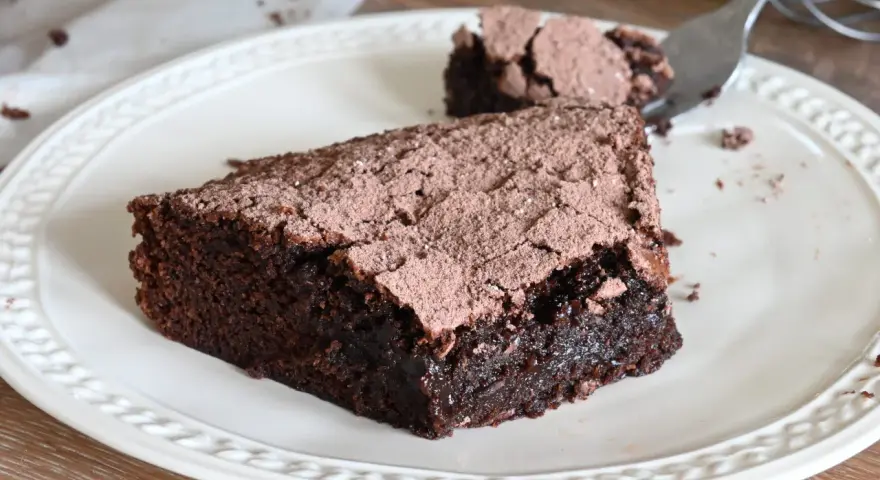 Una fetta di torta al cioccolato fondente, cosparsa di cacao, su un piatto bianco. Un pezzetto è stato staccato con una forchetta.