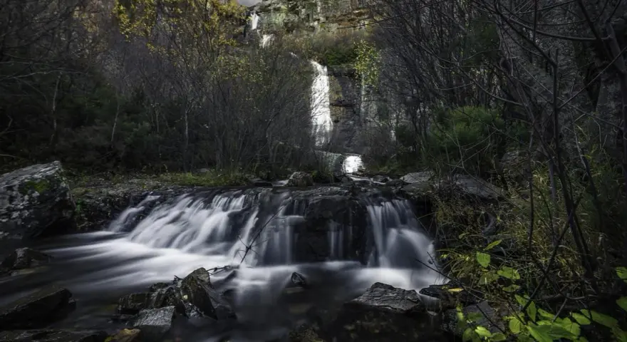 Cascada en el **río Bornova**, con aguas sedosas cayendo entre rocas y vegetación otoñal, bajo un cielo nublado.