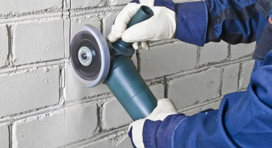 Hands in gloves use an angle grinder to cut a line into a white brick wall, demonstrating how to cut brick precisely.