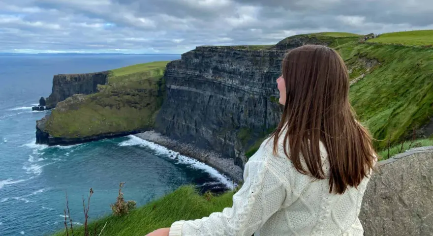 A woman in a cream cable-knit sweater gazes at the dramatic Cliffs of Moher, a breathtaking highlight of many tours of Ireland.