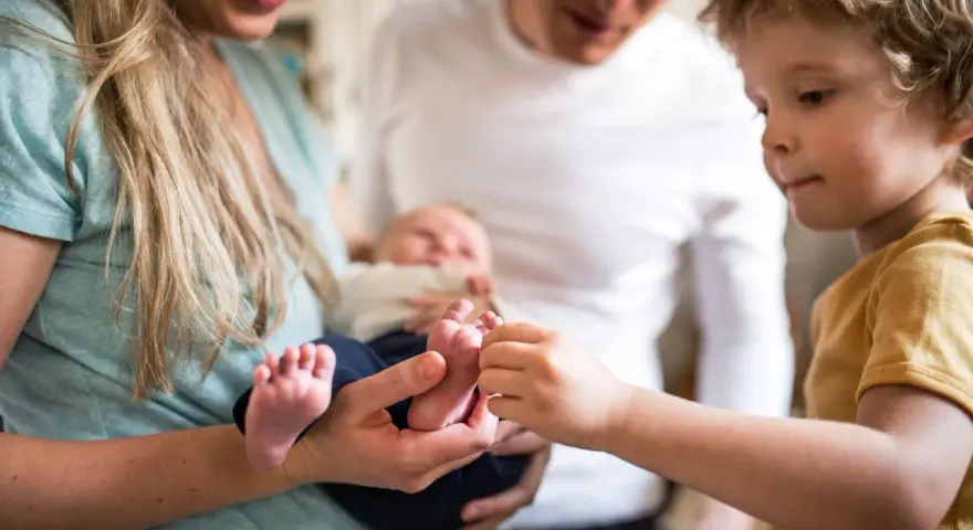 Ein älteres Kind berührt neugierig die Füßchen des Babys. Ein Hauch von eifersucht unter geschwistern liegt in der Luft, während die Eltern das neue Familienmitglied halten.