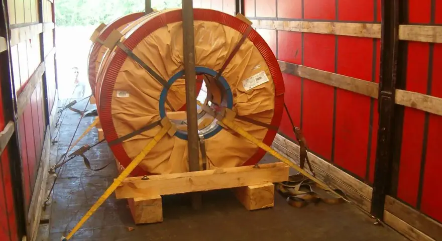 Dwa wielkie zwoje metalu zabezpieczone drewnianym mocowaniem blokowym i pasami transportowymi w naczepie.