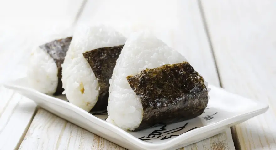 Trzy ryżowe kulki onigiri z nori na białym talerzyku. Idealne na szybki lunch, oto prosty onigiri przepis.