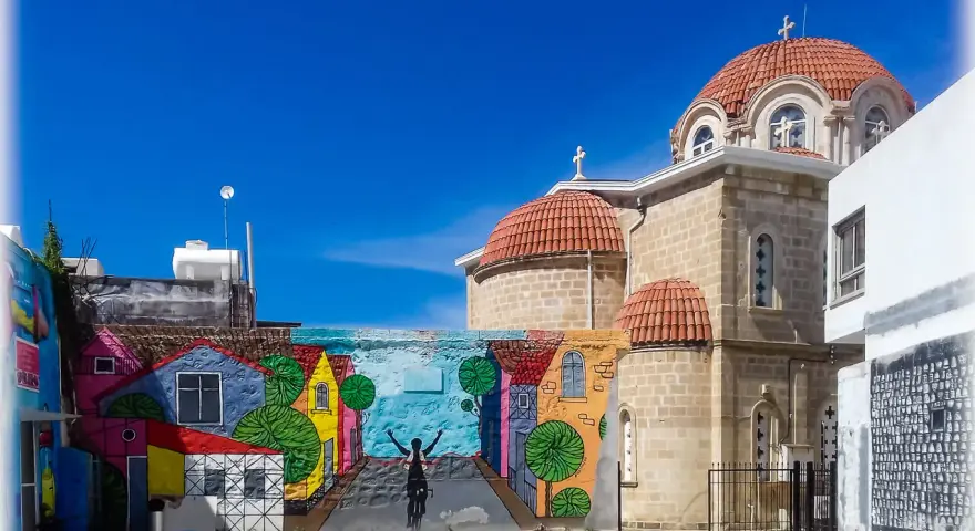 Kolorowy mural z domkami i rowerzystą zdobi ścianę w Pafos, stare miasto. Obok wznosi się kamienna cerkiew z pomarańczowymi kopułami.