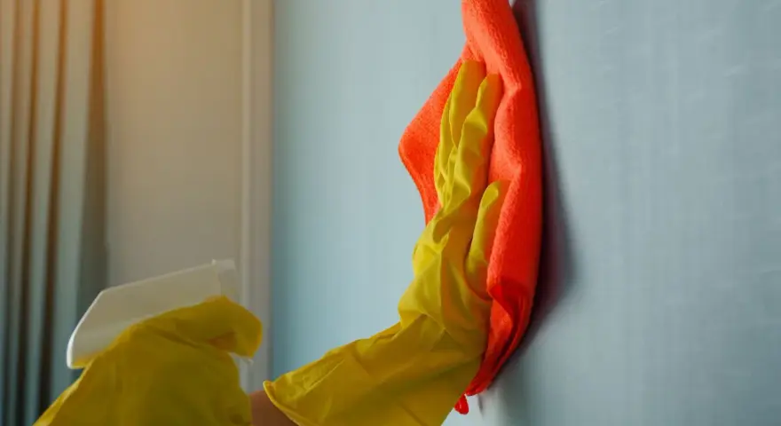 Ręka w żółtej rękawiczce z pomarańczową ściereczką czyści ścianę. Dowiedz się, czym zmyć farbę ceramiczną, by przywrócić jej blask.