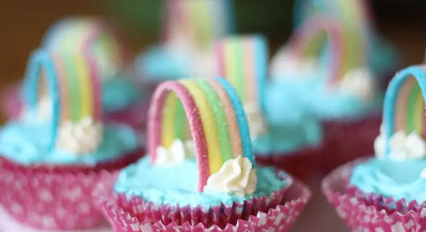 Sju cupcakes med ljusblå regnbågs frosting och godisregnbågar. Rosa muffinsformar med mönster.