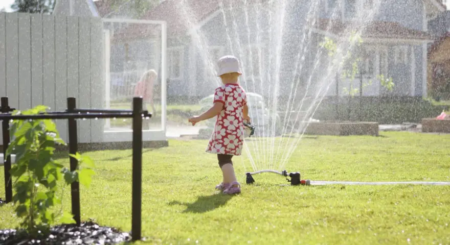 Lekfull **tipspromenad barn** i sommarregn från en vattenspridare. Ett litet barn i blommig klänning njuter av vattnet på gräsmattan.