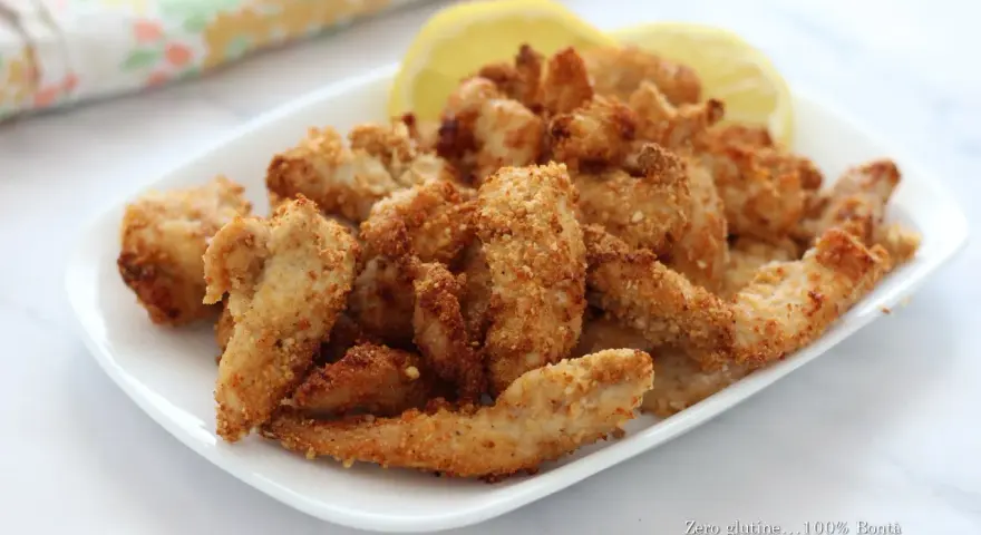 Straccetti di pollo in friggitrice ad aria, dorati e croccanti, serviti su un piatto bianco con spicchi di limone.