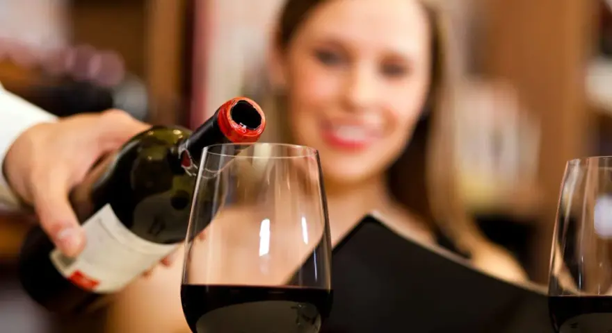 Kelner nalewa wino, a uśmiechnięta kobieta czeka na kieliszek. Wino zawiera siarczyny, które pomagają w jego konserwacji.