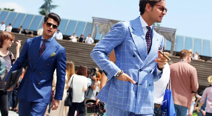 Dwóch eleganckich mężczyzn w garniturach prezentuje letnią modę męską. Jeden w niebieskim, drugi w błękitnym w kratę, obaj z pewnością siebie.