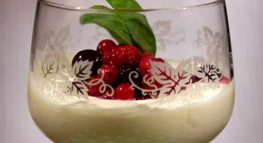 En elegant glas med vit chokladmousse, toppad med färska bär och ett grönt blad. Glaset har en vacker dekor av löv.