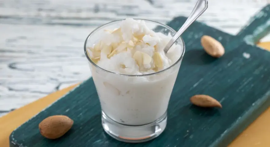 Rinfrescante granita alla mandorla, guarnita con lamelle di mandorle, servita in un bicchiere su un tagliere scuro.
