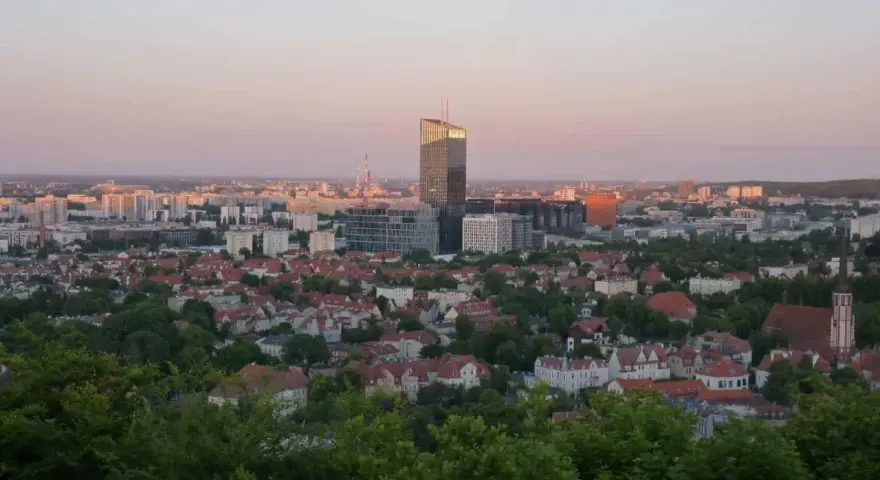 Widok na miasto o zachodzie słońca. W oddali widać nowoczesne budynki, a bliżej dachy domów i zieleń. Jakby pachołek oliwa na tle nieba.