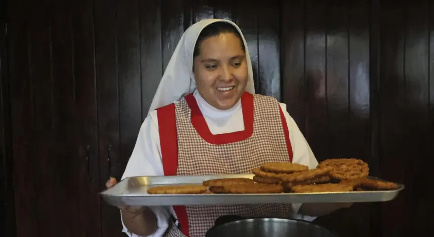 A smiling nun in a white habit and checkered apron holds a tray of fried pastries, perhaps answering the question: what do nuns eat for breakfast?
