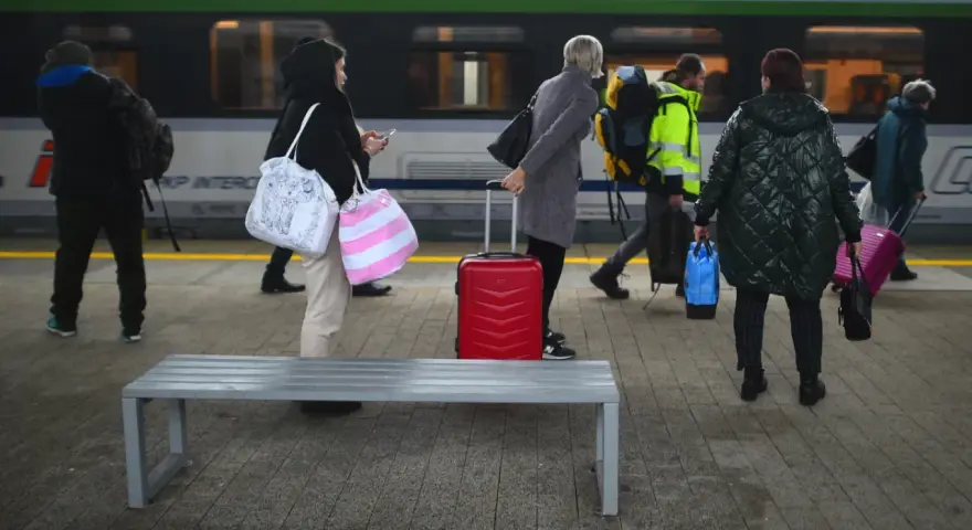 Ludzie z bagażami czekają na peronie PKP Intercity. Czerwona walizka stoi obok ławki.