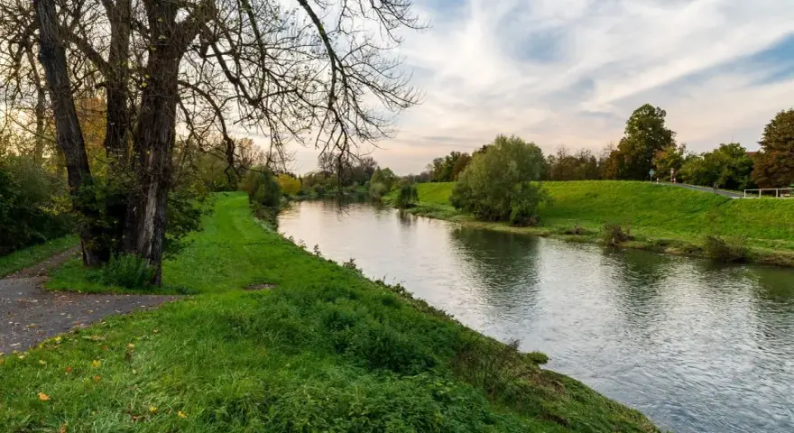 Spokojna rzeka na u, otoczona zielonymi brzegami i drzewami. W oddali widać ścieżkę i niebo z chmurami.