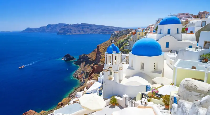 Santorini wycieczki: białe domy z niebieskimi kopułami na klifie nad błękitnym morzem. Statek płynie w oddali.